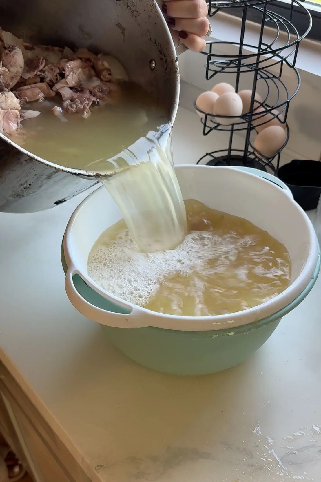 Straining homemade chicken stock through a colander to remove bones and large ingredients, resulting in a clear, flavorful broth.